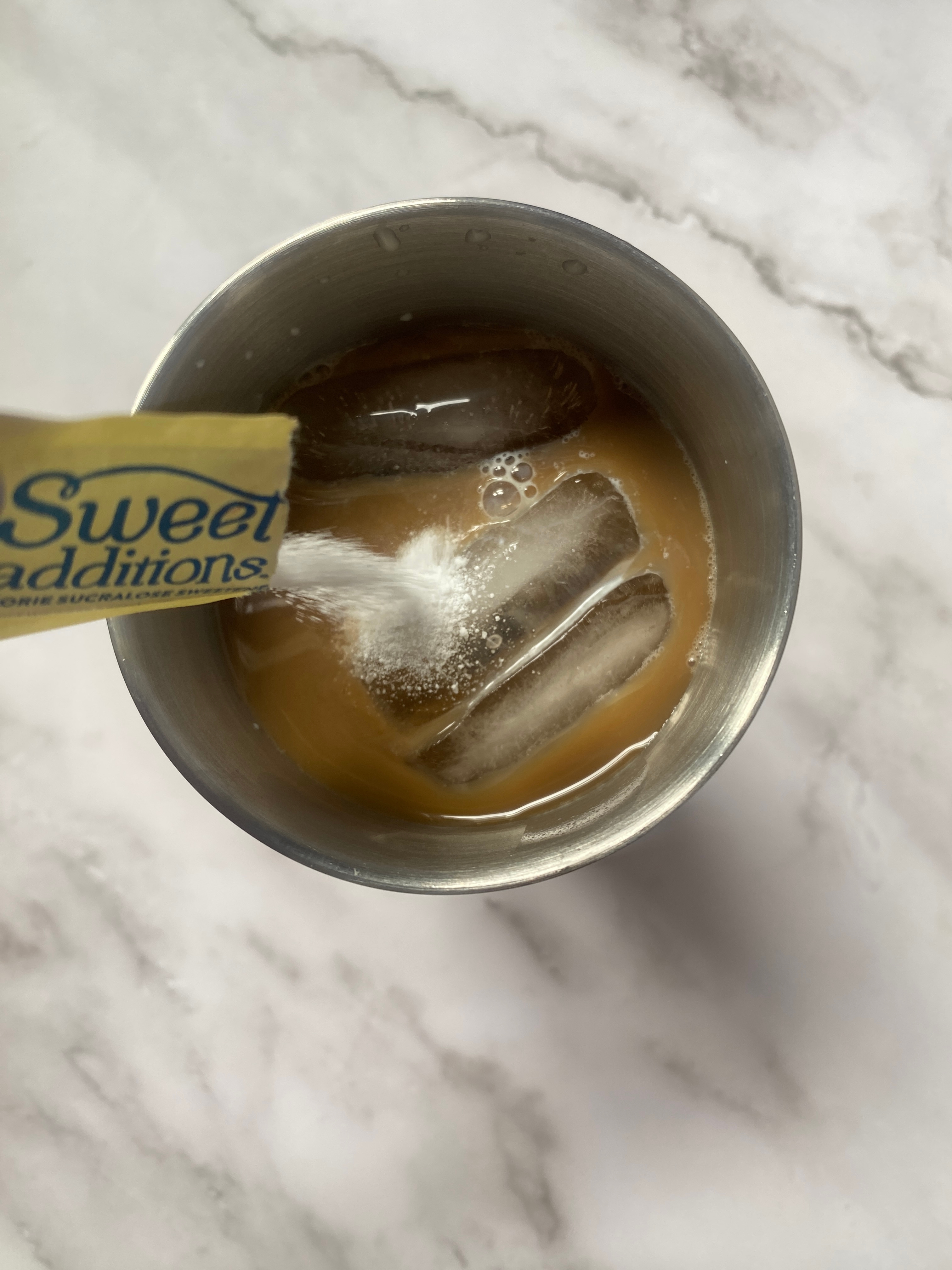 Sweetener being added to iced coffee in a stainless steel cocktail shaker on a marble surface