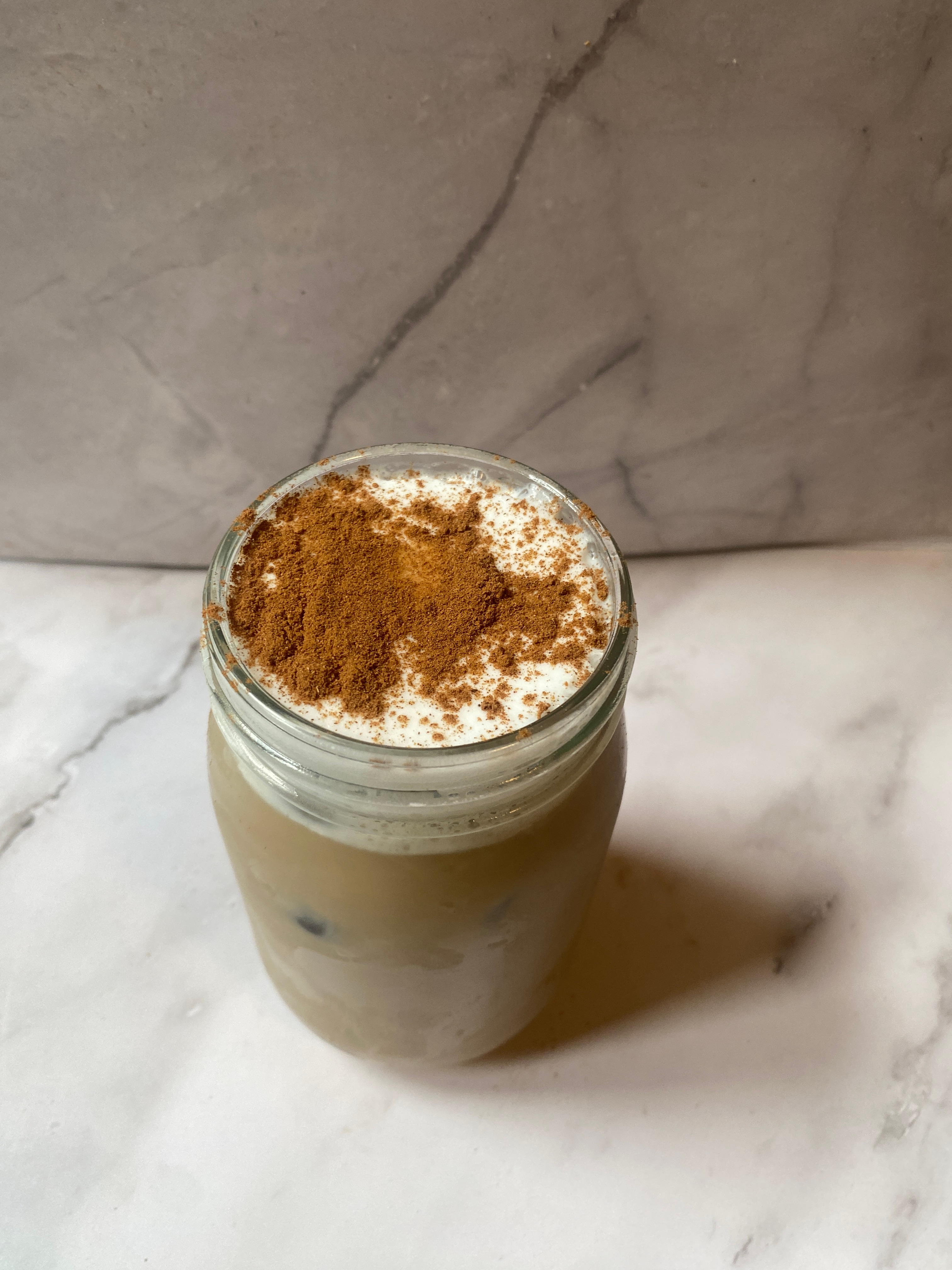 Iced coffee in a glass jar topped with foam and cinnamon on a marble surface