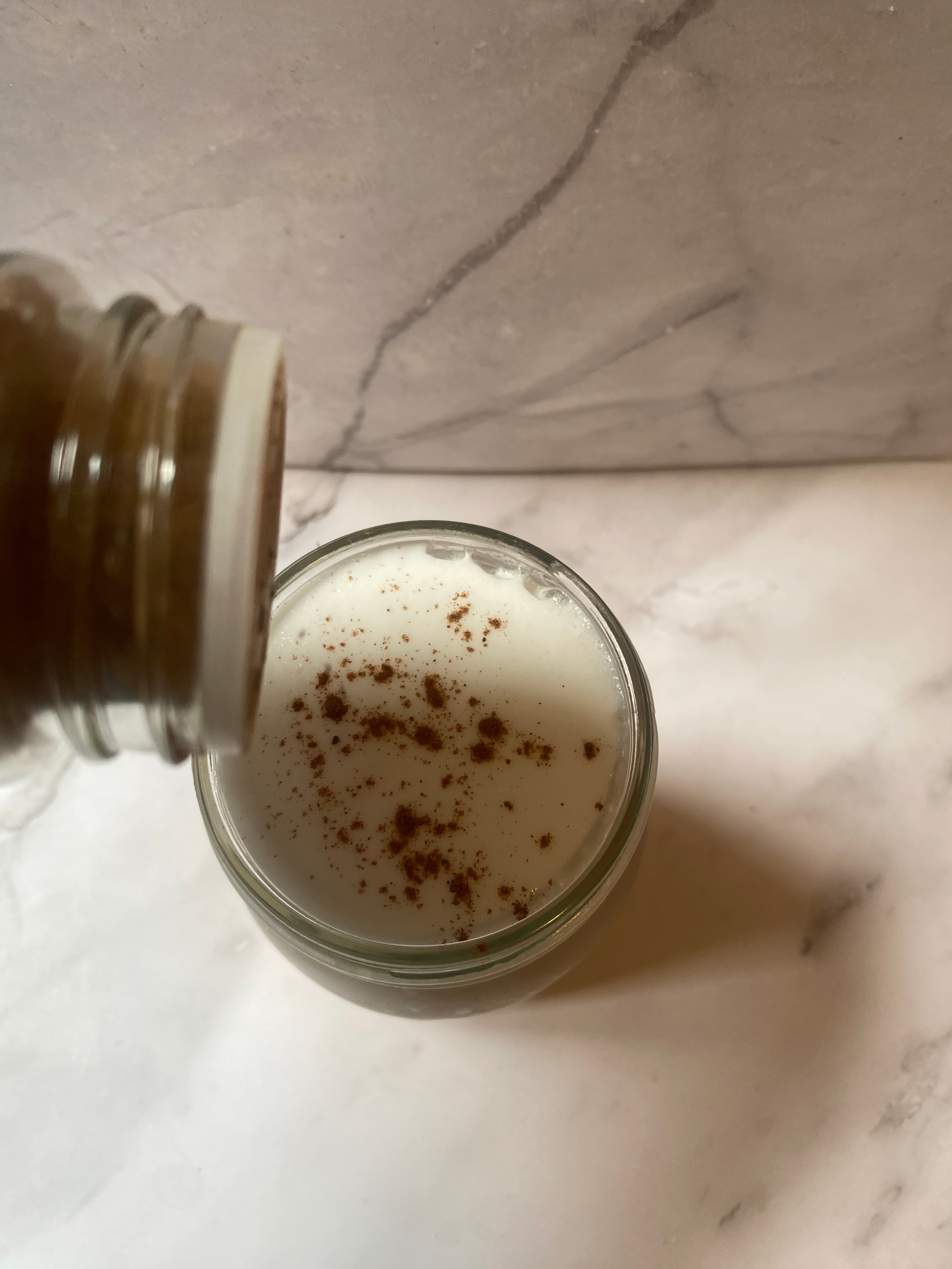 A jar pouring cinnamon spice onto a glass of frothy milk on a marble countertop