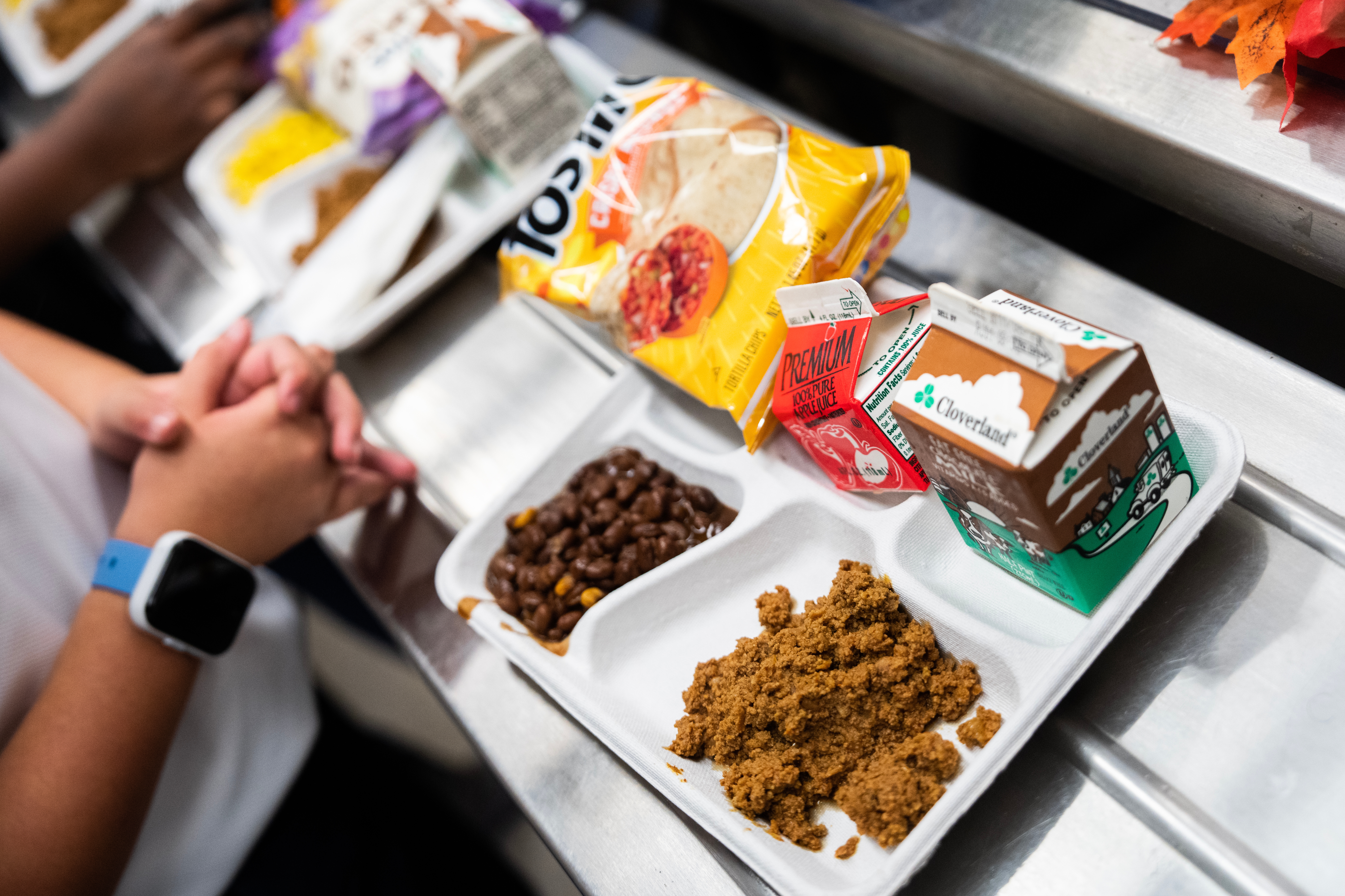 A tray with beans, tortilla chips, and two milk cartons on a metal surface. Hands clasped nearby, suggesting a school lunch setting