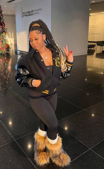 Person successful  a stylish varsity jacket, achromatic  leggings, and fur boots posing indoors, flashing a bid    sign