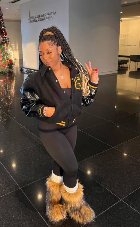 Person successful a stylish varsity jacket, achromatic leggings, and fur boots posing indoors, flashing a bid sign