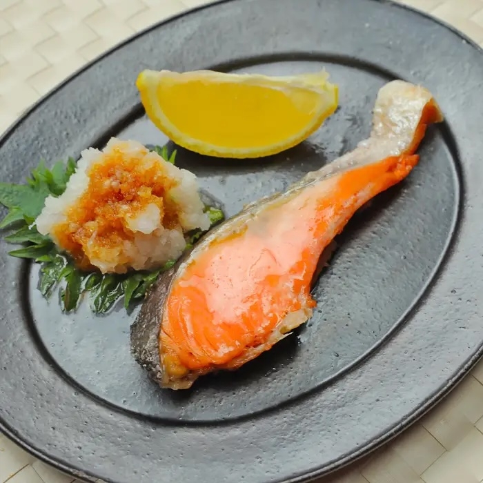 焼き鮭、大根おろしとレモンが黒い皿に盛られた料理。食材が整然と配置されている。