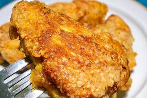 フォークで持ち上げられたジューシーなフライドチキンのクローズアップショット。後ろに白いプレートに盛られたチキンがある。