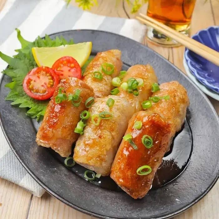 皿に盛り付けられたミニトマト添えの豚肉巻き。青ネギがかかり、横にレモンと大葉もあります。シンプルな食事風景。