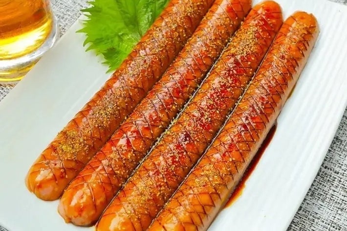 4本のカットされた焼きソーセージが皿に縦に並び、横に緑の葉と飲み物がある。
