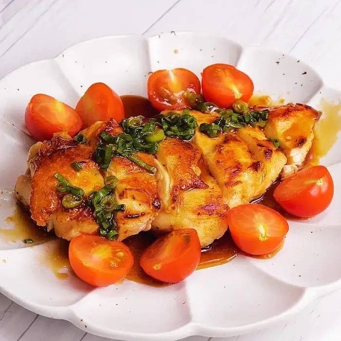 白い皿に盛られた焼きチキン、ネギとソースがかかり、周りに切ったミニトマトが配置されています。