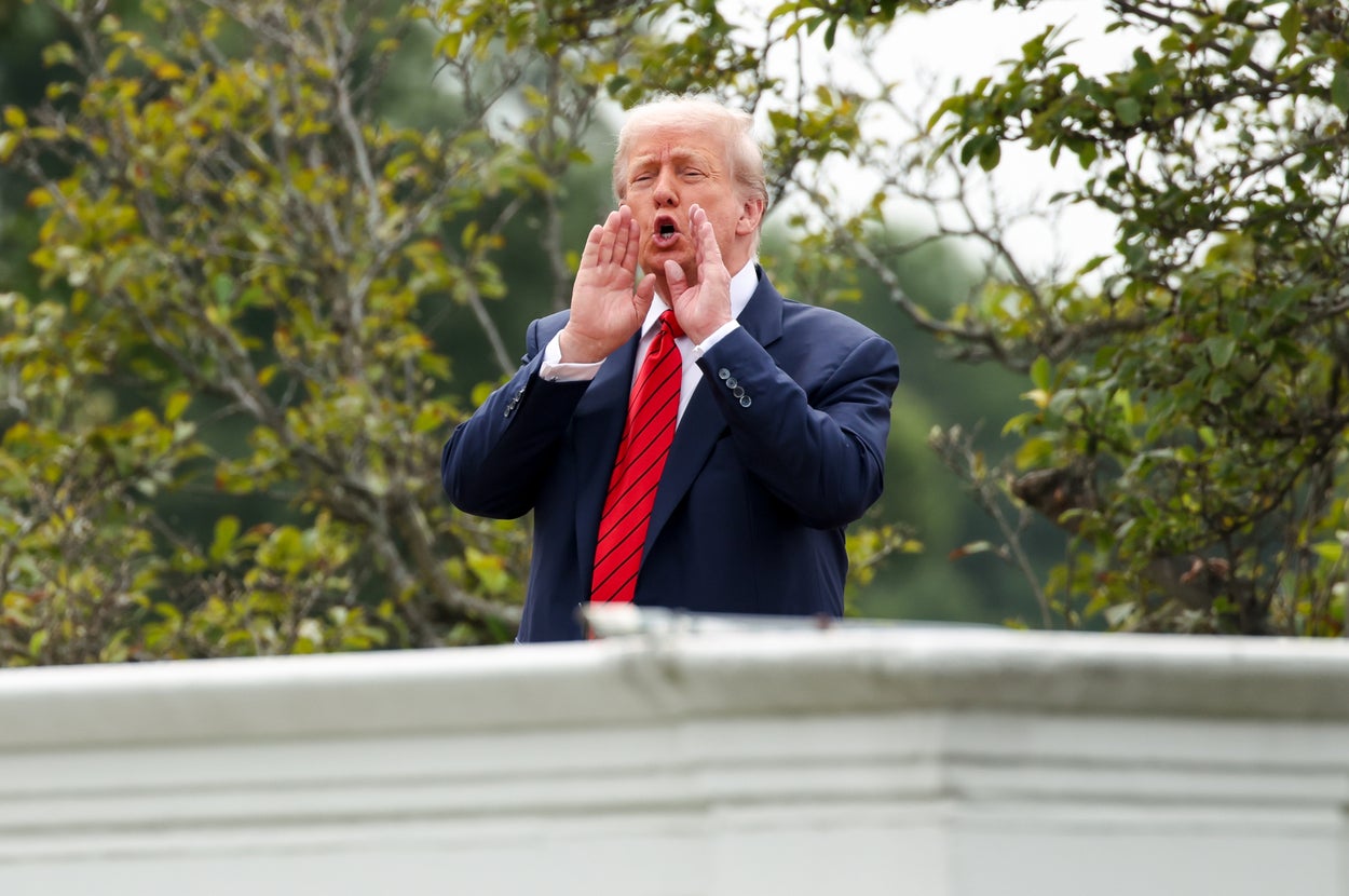 Person outdoors, wearing a suit with a red tie, cupping hands around mouth to shout or call out. Trees are visible in the background