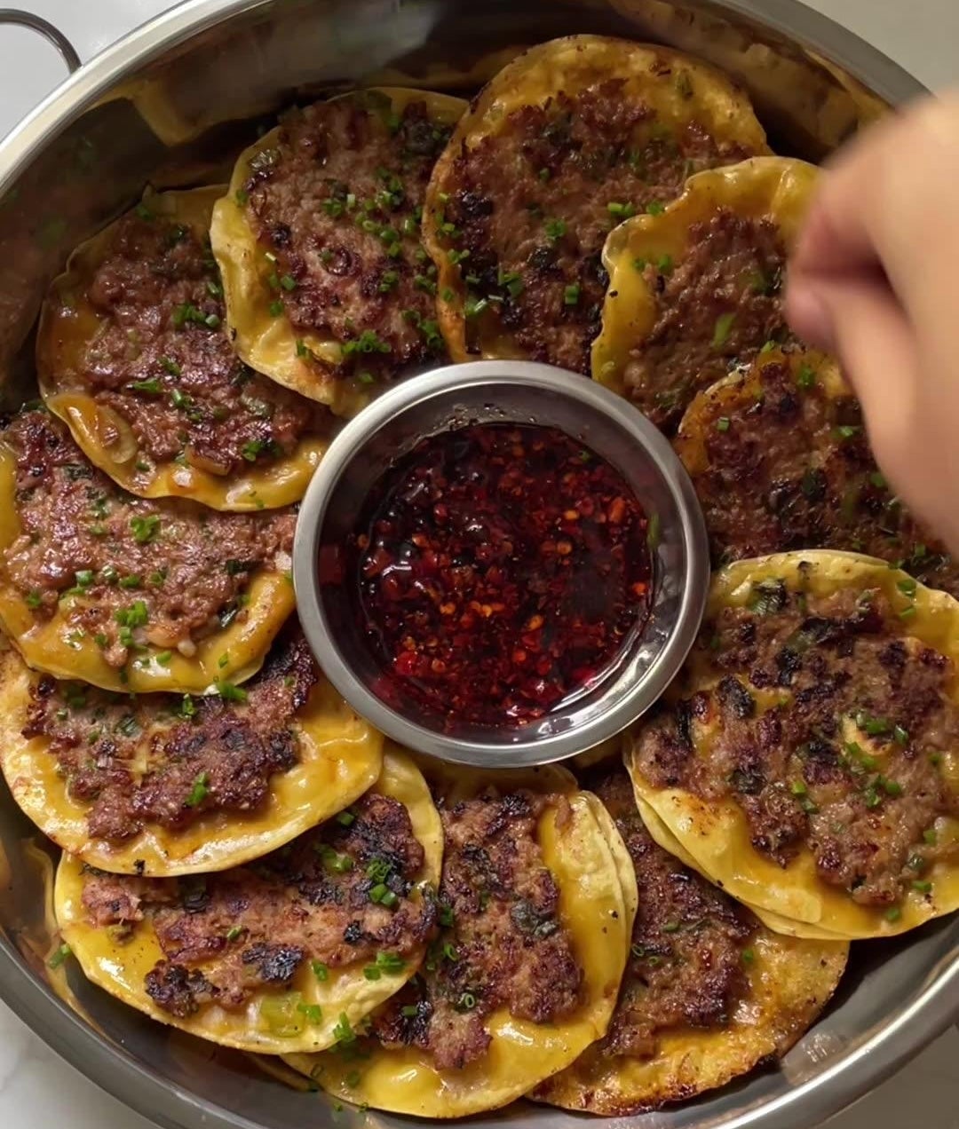 A platter of round, pan-fried dumplings with a beef filling and a side of chili oil in the center. A hand reaches toward the dish