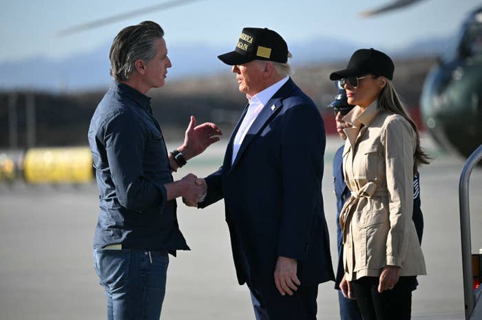 Two men, one in a cap and suit, shake hands outdoors on a tarmac. A woman in a trench coat and cap stands nearby. Helicopter in background