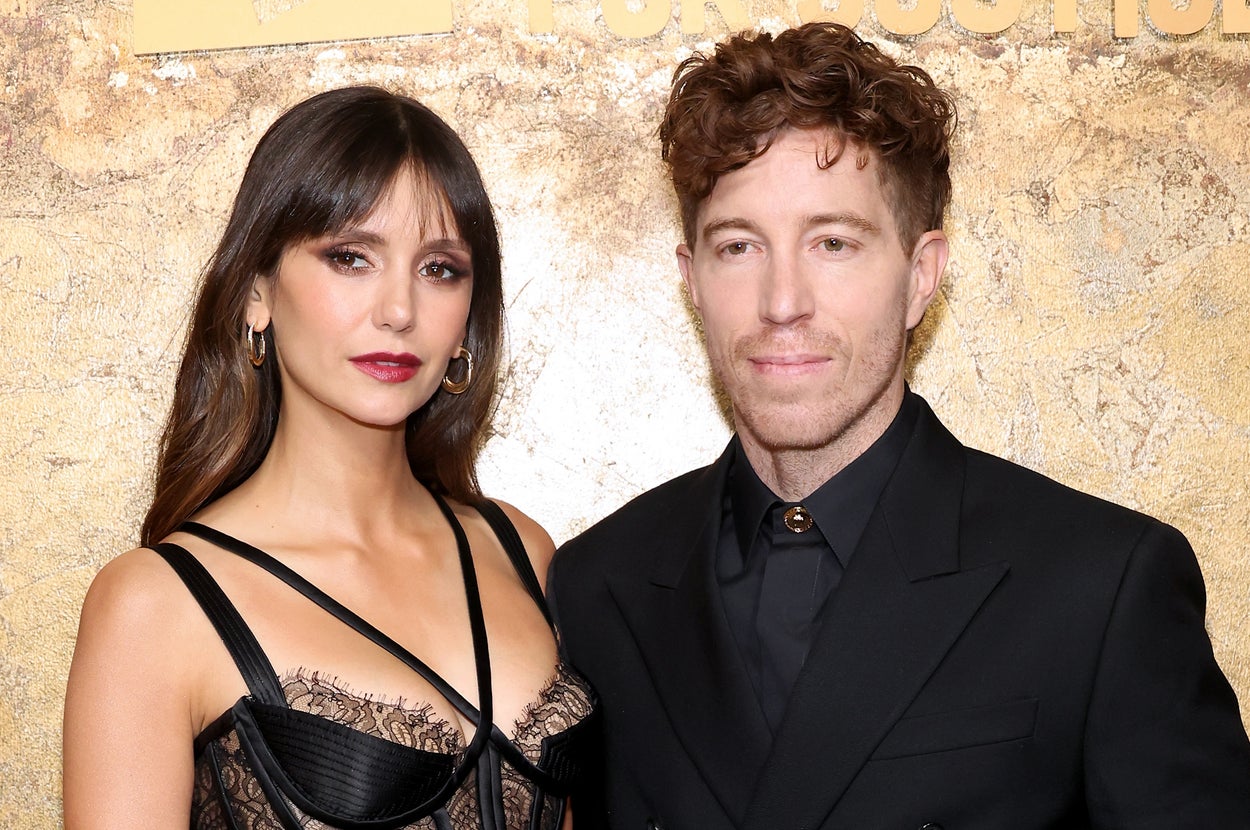 Two celebrities posing together; woman in a stylish lace corset top, man in a tailored dark suit. Ornate gold backdrop