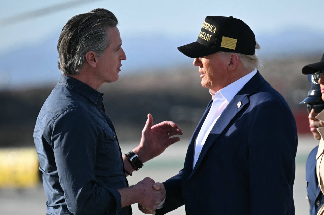 Two men in casual attire shake hands at an outdoor event, both wearing dark clothes. One wears a cap with visible writing