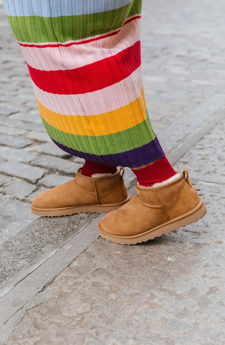 Person wearing brown shearling slippers and rainbow-striped pants walks on cobblestone