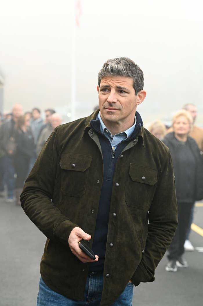 Man in a casual jacket holding a phone, standing outside with a misty background and people in the distance