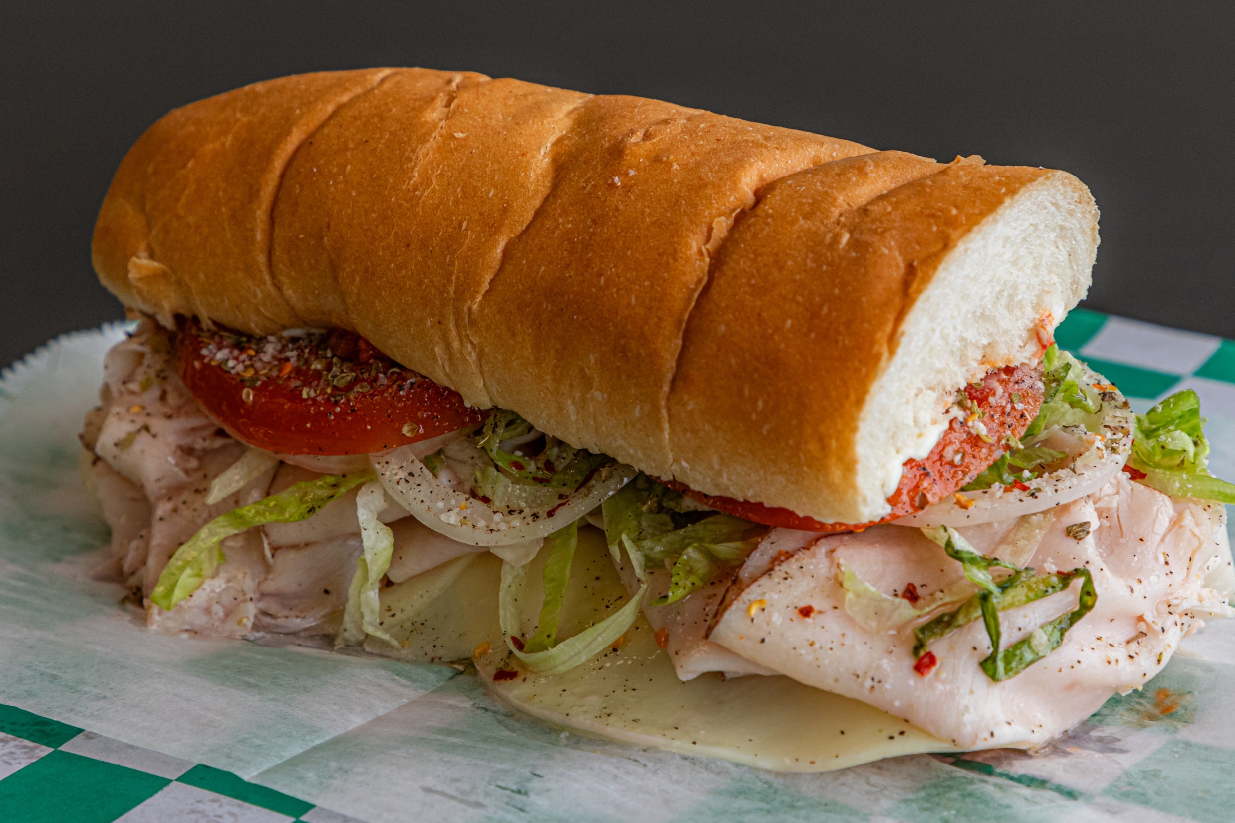 A freshly made sandwich on a sub roll stacked with turkey, provolone cheese, lettuce, tomato, onion, and seasoning on a checkered paper wrapper