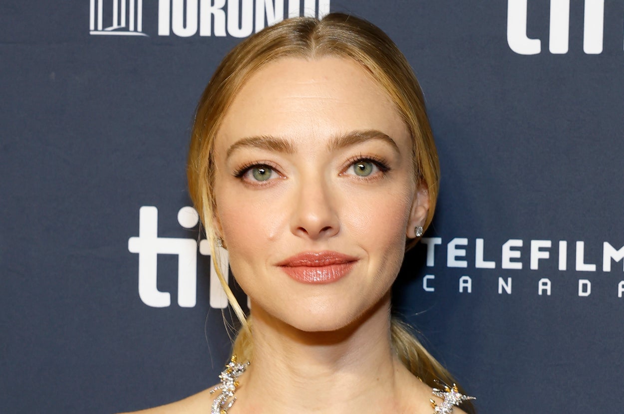 Person at film event wearing an off-shoulder dress with floral embellishments and a decorative necklace, in front of a branded backdrop