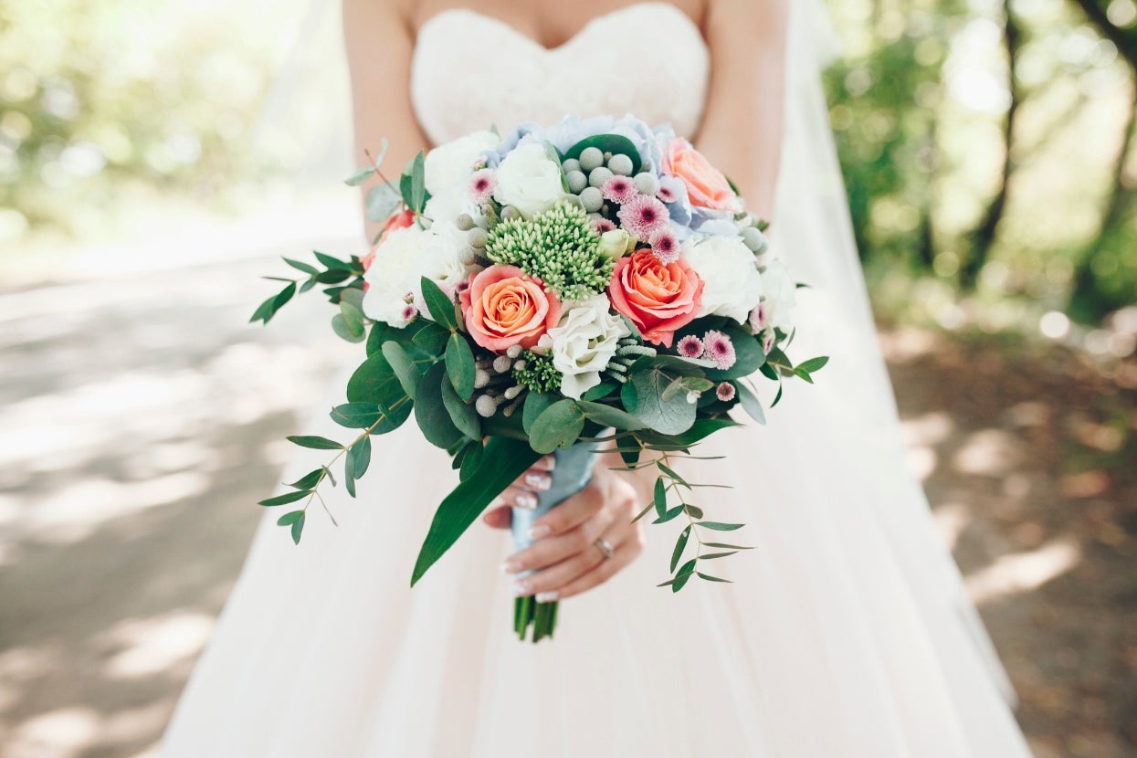 Bride holding a bouquet of mixed flowers, including roses and greenery, outdoors in a natural setting