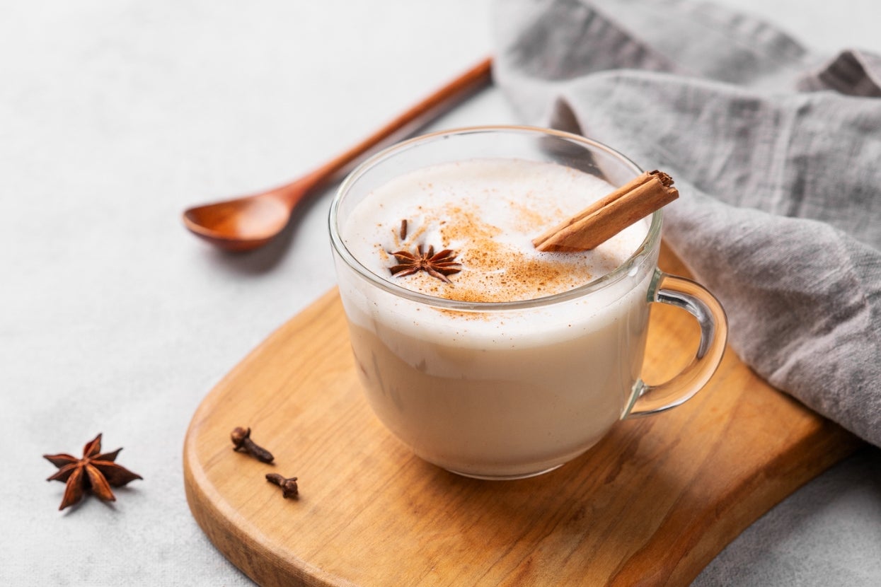 A glass cup of creamy chai tea topped with cinnamon sticks and star anise, set on a wooden board with a linen cloth and wooden spoon nearby