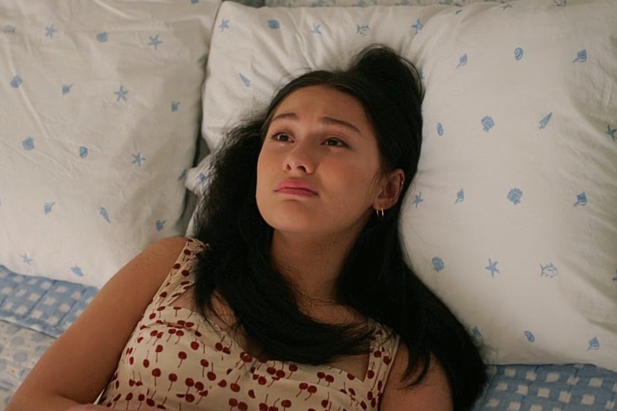 A young woman with long hair, wearing a patterned top, lies on a bed looking contemplative