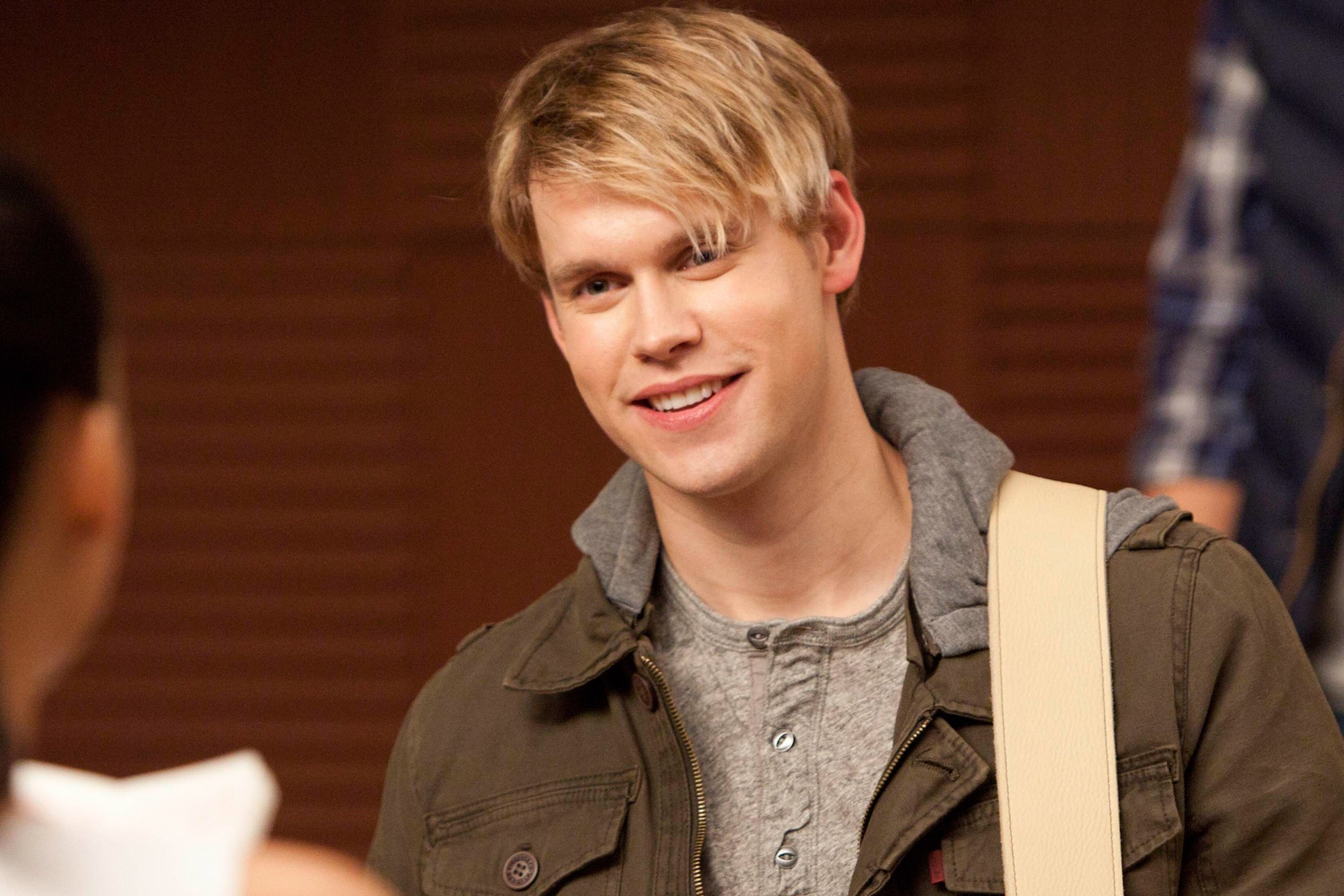 Person playing guitar, wearing casual layered clothing, smiles at someone in a cheerleader uniform indoors