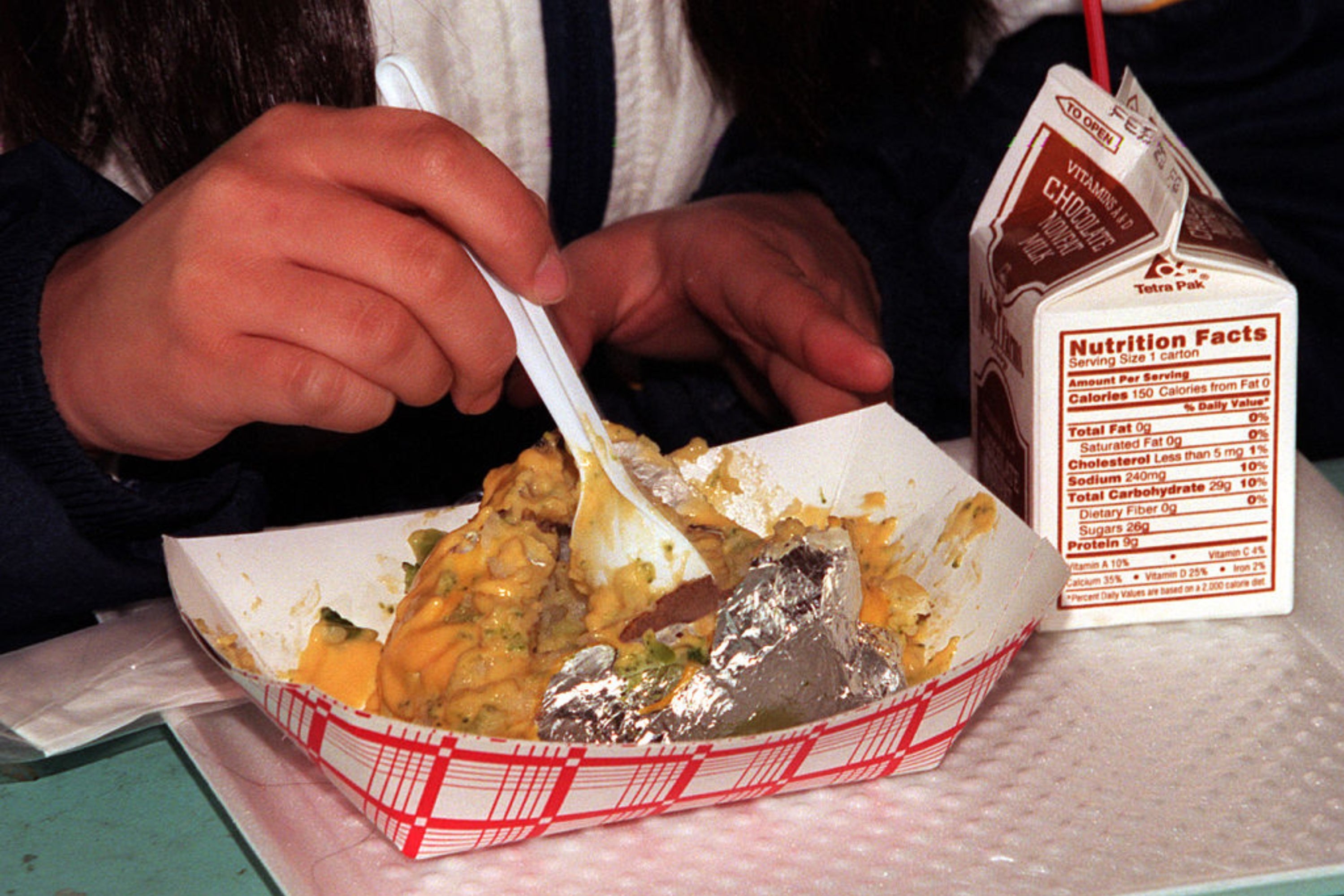 Person eating a baked potato topped with melted cheese and broccoli from a disposable tray using a plastic fork, next to a carton of chocolate milk
