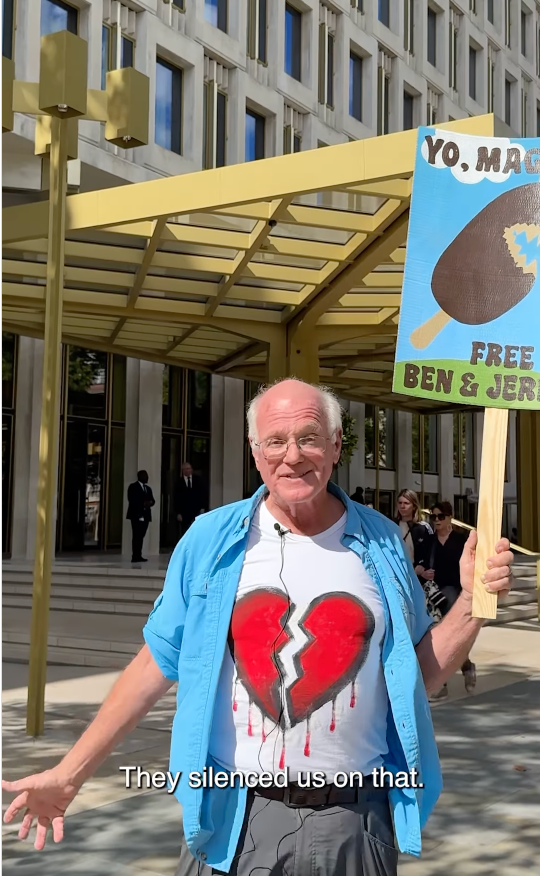 Man holding protest sign stands in front of a building. He's wearing a T-shirt with a broken heart design. Subtitles read: "They silenced us on that."