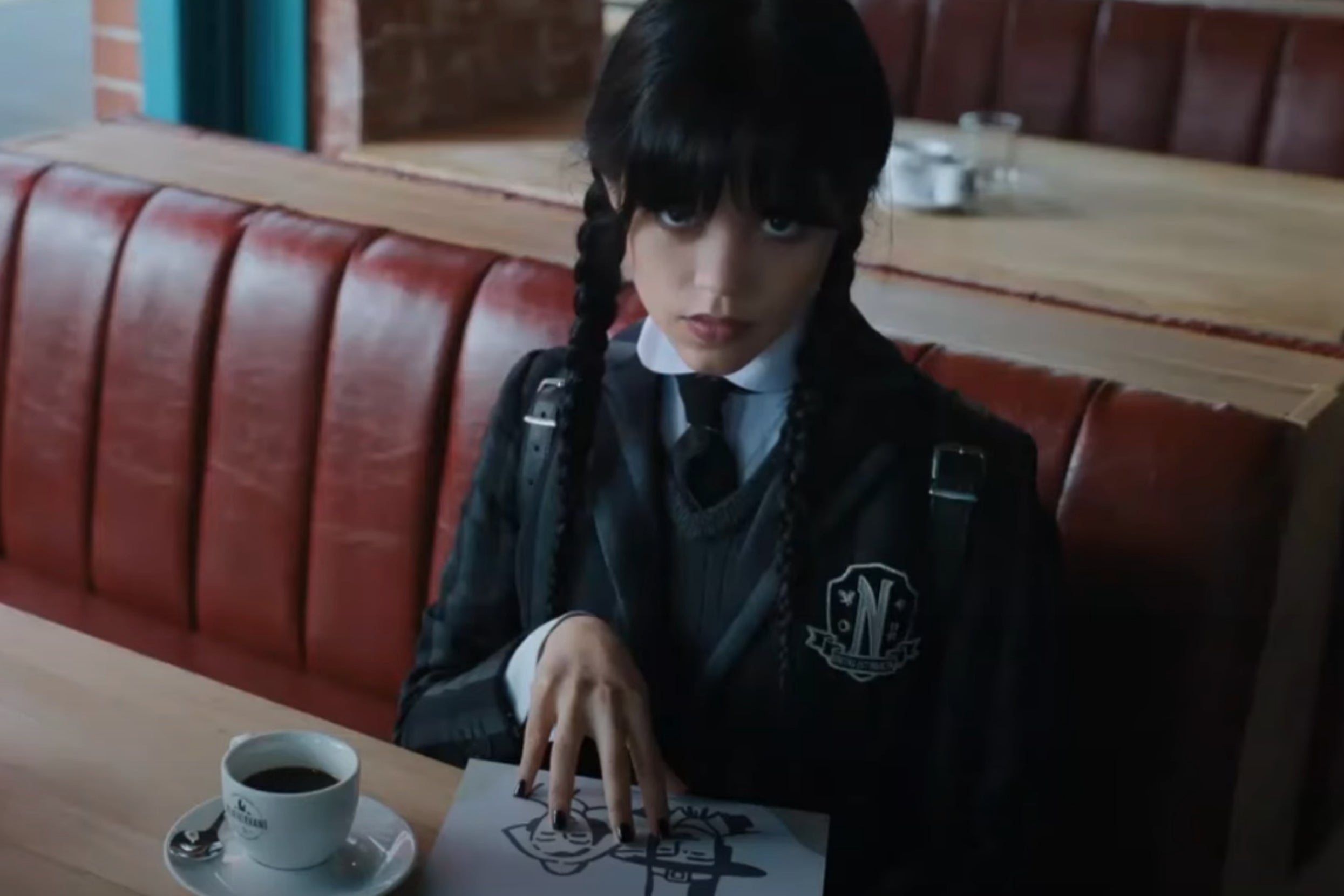 Wednesday Addams sits at a diner booth, with a cup of coffee and a paper with drawings on the table