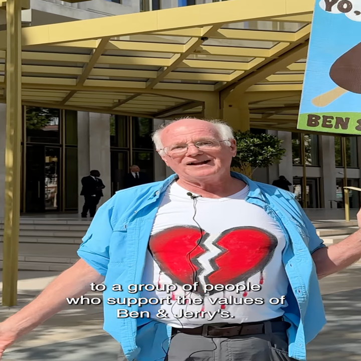 Man in shirt with broken heart graphic, holding a pro-Ben &amp; Jerry's sign outside a building, speaking to a group