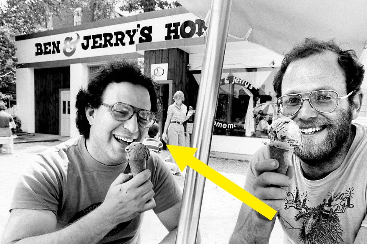 Two men smiling, holding ice cream cones in front of a Ben & Jerry's store. A yellow arrow points to a woman behind them