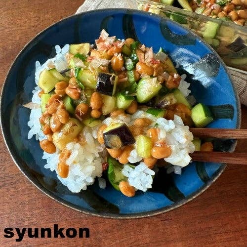 青い皿に盛られたご飯の上に、納豆、キュウリ、ナスが乗っている料理。食卓に木製の箸が添えられている。