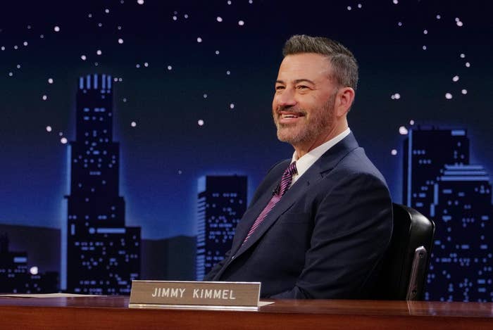 Two radical are seated connected a speech amusement set, smiling and talking. One is wearing a velvet suit, and the different is successful a suit down a desk