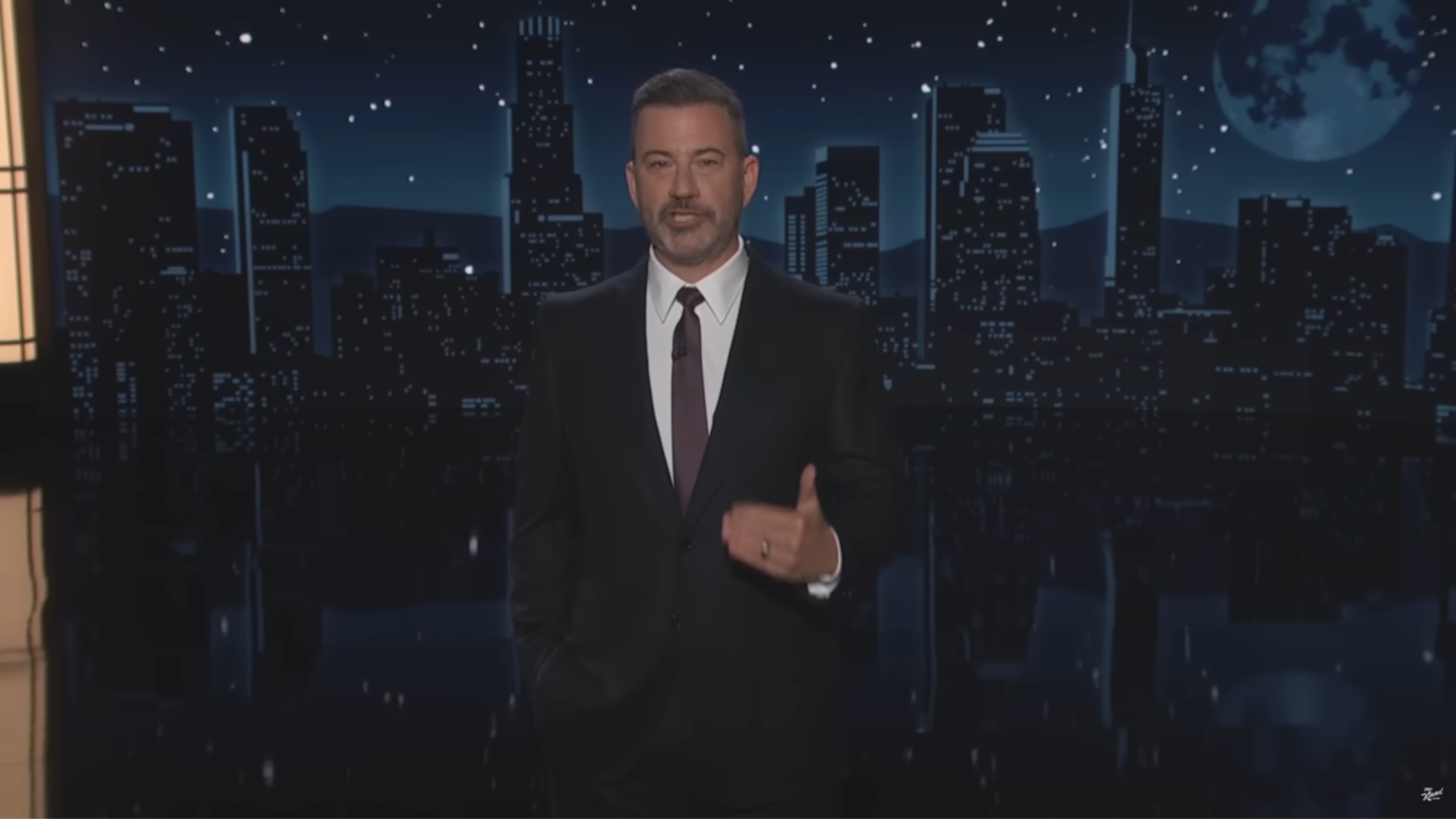 A person in a suit stands on a stage with a city skyline backdrop at night