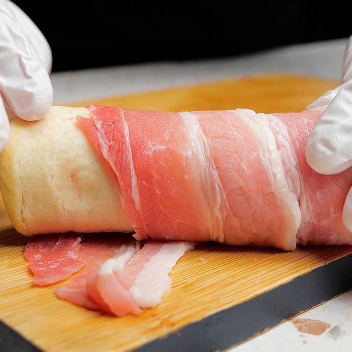 手袋をした手が、薄く切った生ハムで包まれた食材を木製の板の上で巻いている様子。