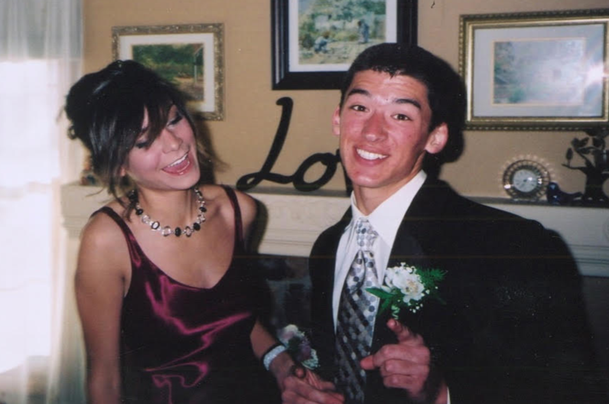 Two people at an event; a woman in a satin dress and a man in a suit with a boutonniere, both smiling and posing for the camera