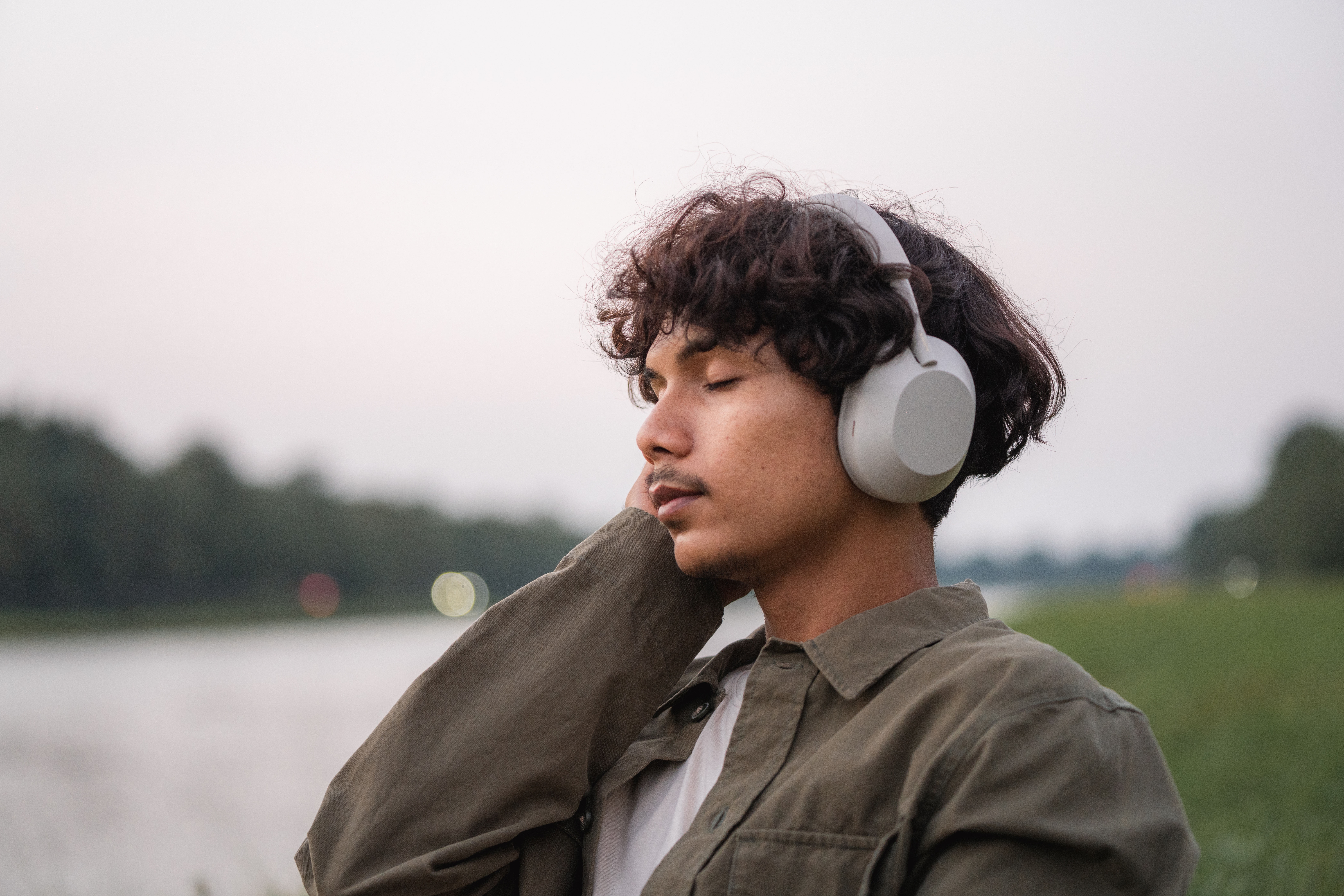 Person med hodetelefoner som sitter utendørs, lukkede øyne, virker avslappet i nærheten av en vannmasse