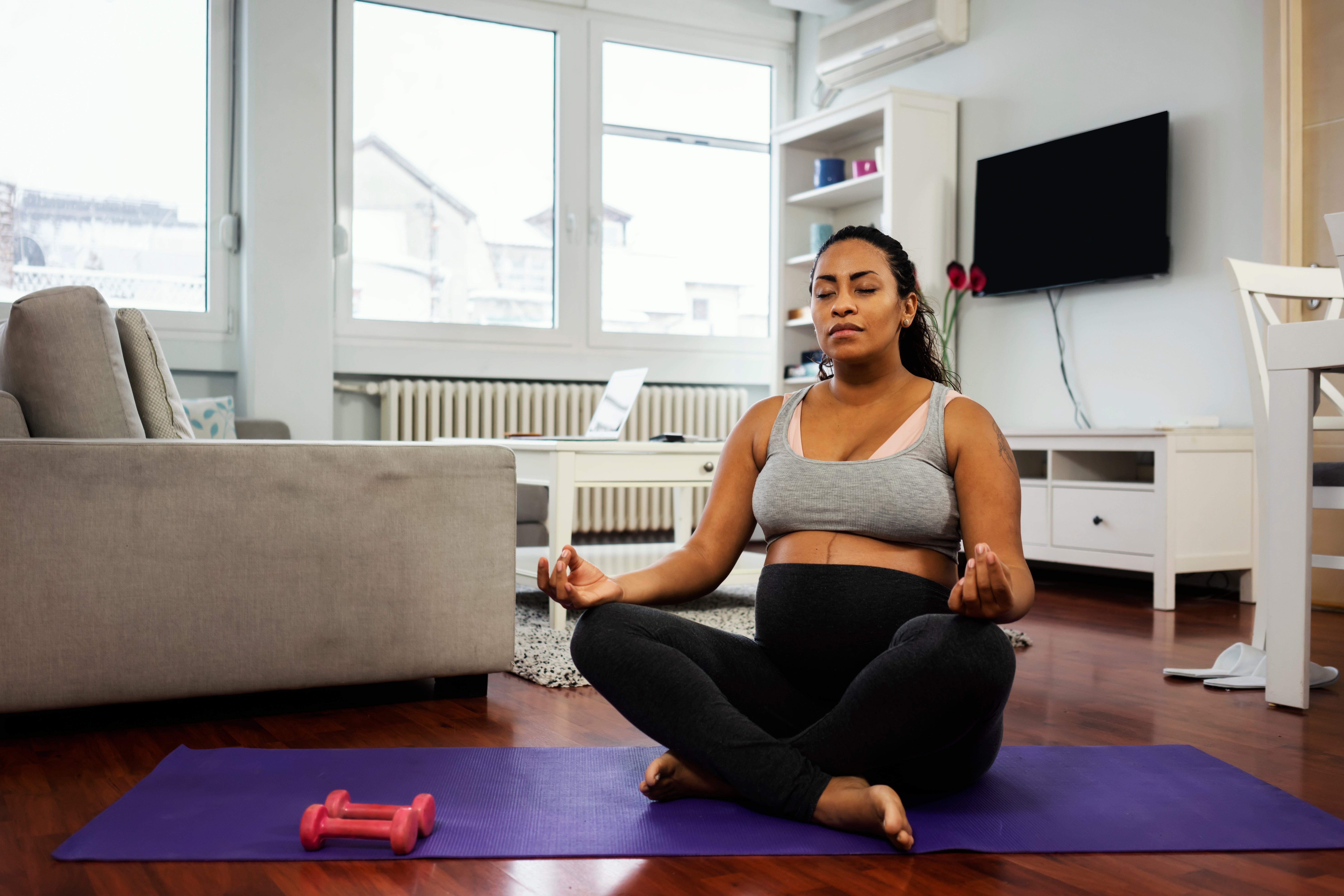 Gravid kvinne mediterer på en yogamatte i en stue, med små hantler i nærheten, og fremmer avslapning og velvære under graviditet