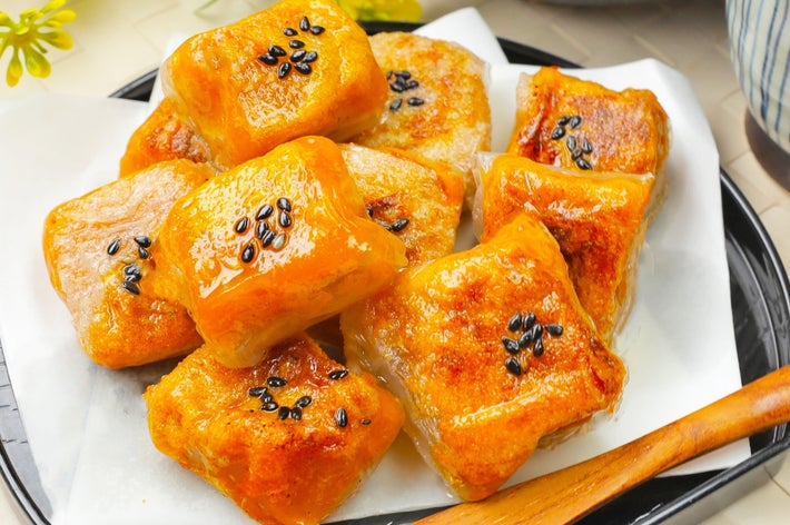 皿に盛られた焼かれたもち菓子に黒いゴマがトッピングされている。木製フォークが添えられている。