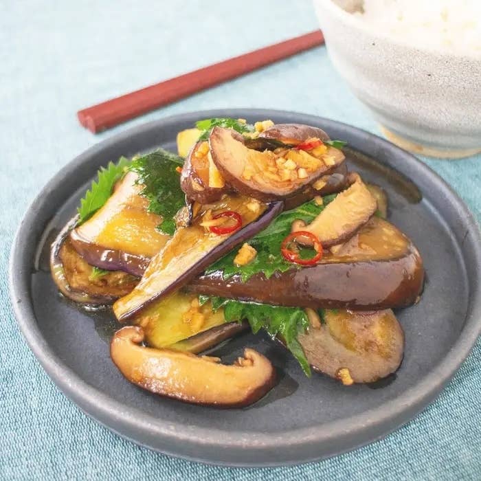 ナスとシイタケの炒め物が盛られた皿。薬味にネギと唐辛子が添えられている。