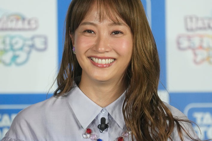 女性がインタビュー会場で微笑んでいます。フリルのあるシャツとカラフルなネックレスを着用しています。