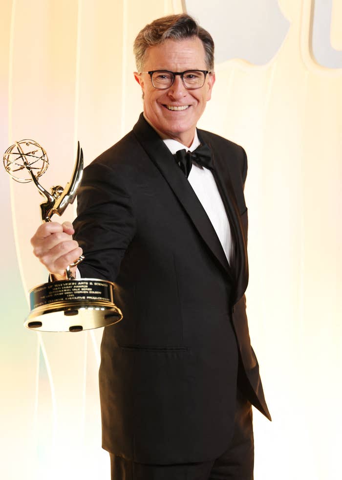 Person in a tuxedo smiles, holding an Emmy award in a celebratory pose at an event