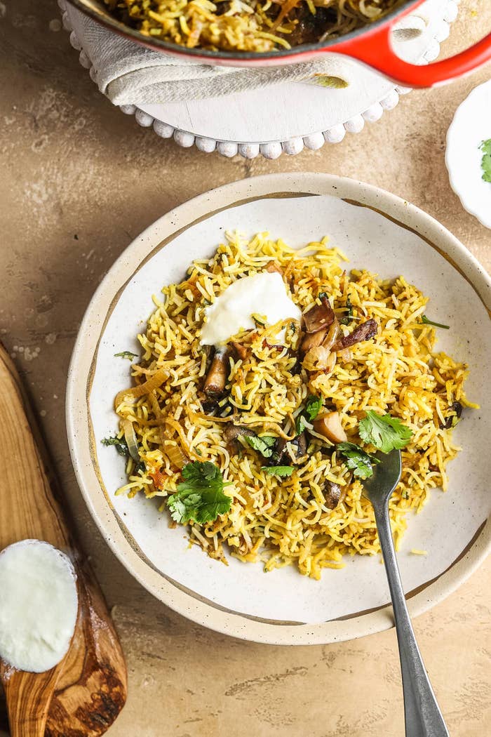 A bowl of biryani with mushrooms, rice, herbs, and a dollop of yogurt on top, placed on a table setting with a fork