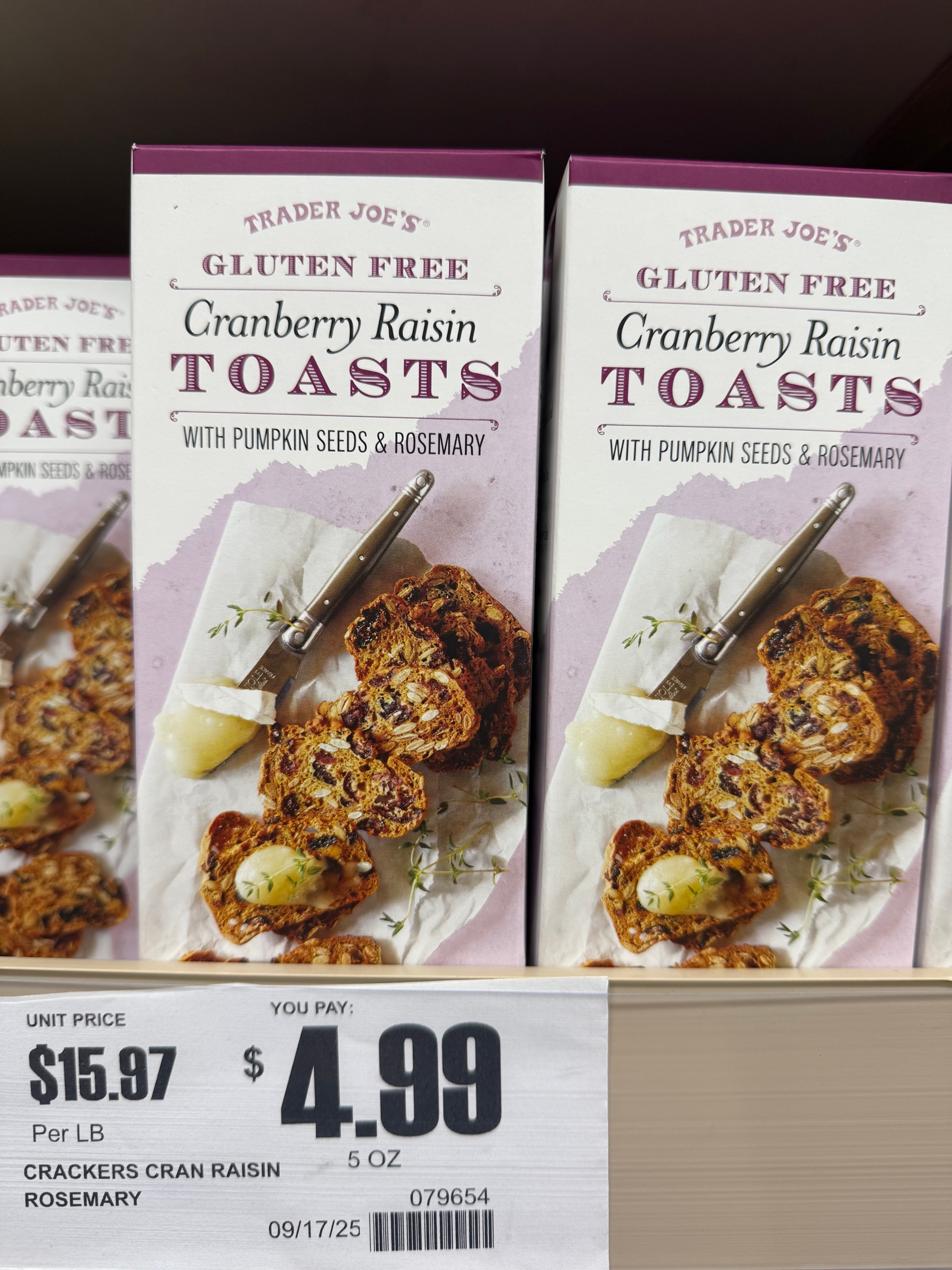Boxes of Trader Joe's Gluten Free Cranberry Raisin Toasts with pumpkin seeds and rosemary displayed on a shelf, priced at $4.99