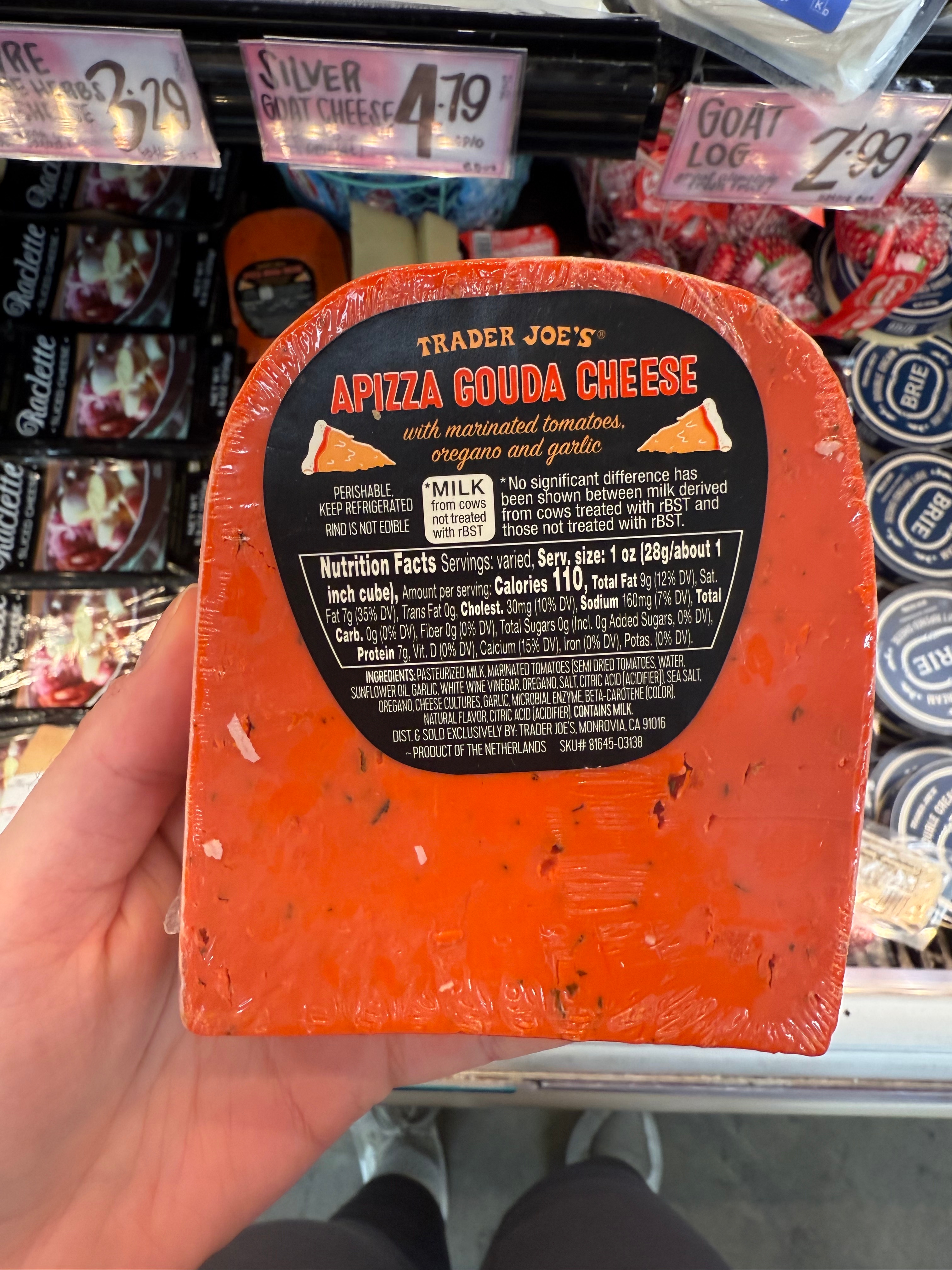A person holds a Trader Joe's Apizza Gouda Cheese with marinated tomatoes, oregano, and garlic. The label includes nutritional information