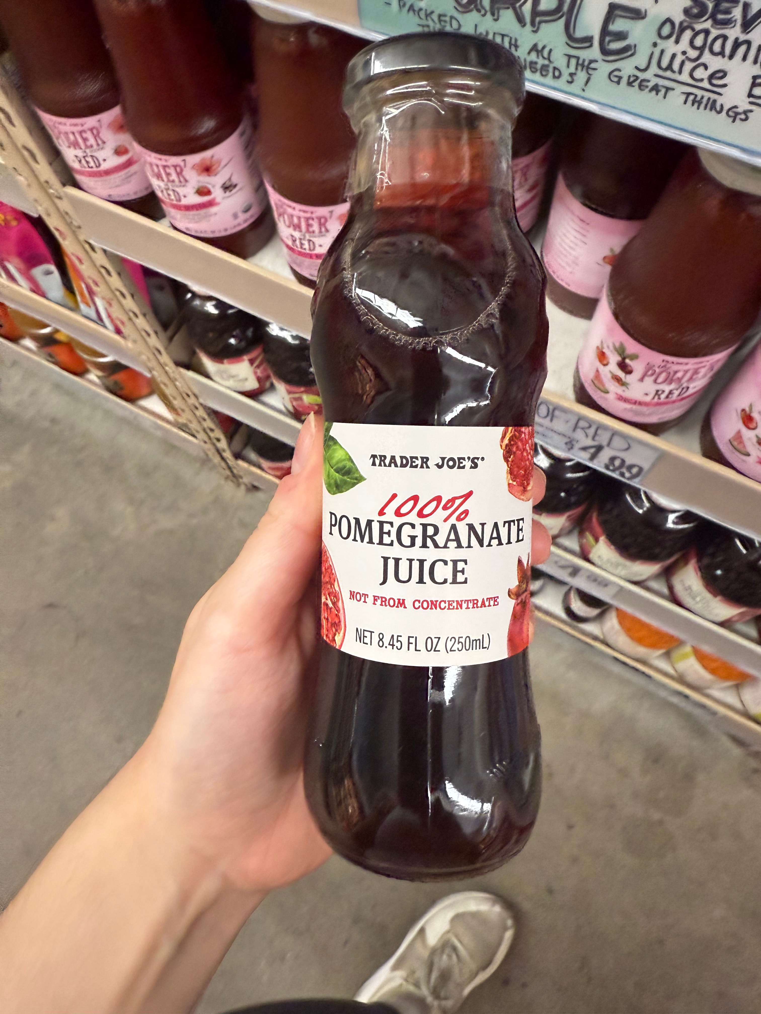 A hand holds a bottle of Trader Joe's 100% Pomegranate Juice in a store aisle with various juice options on the shelves