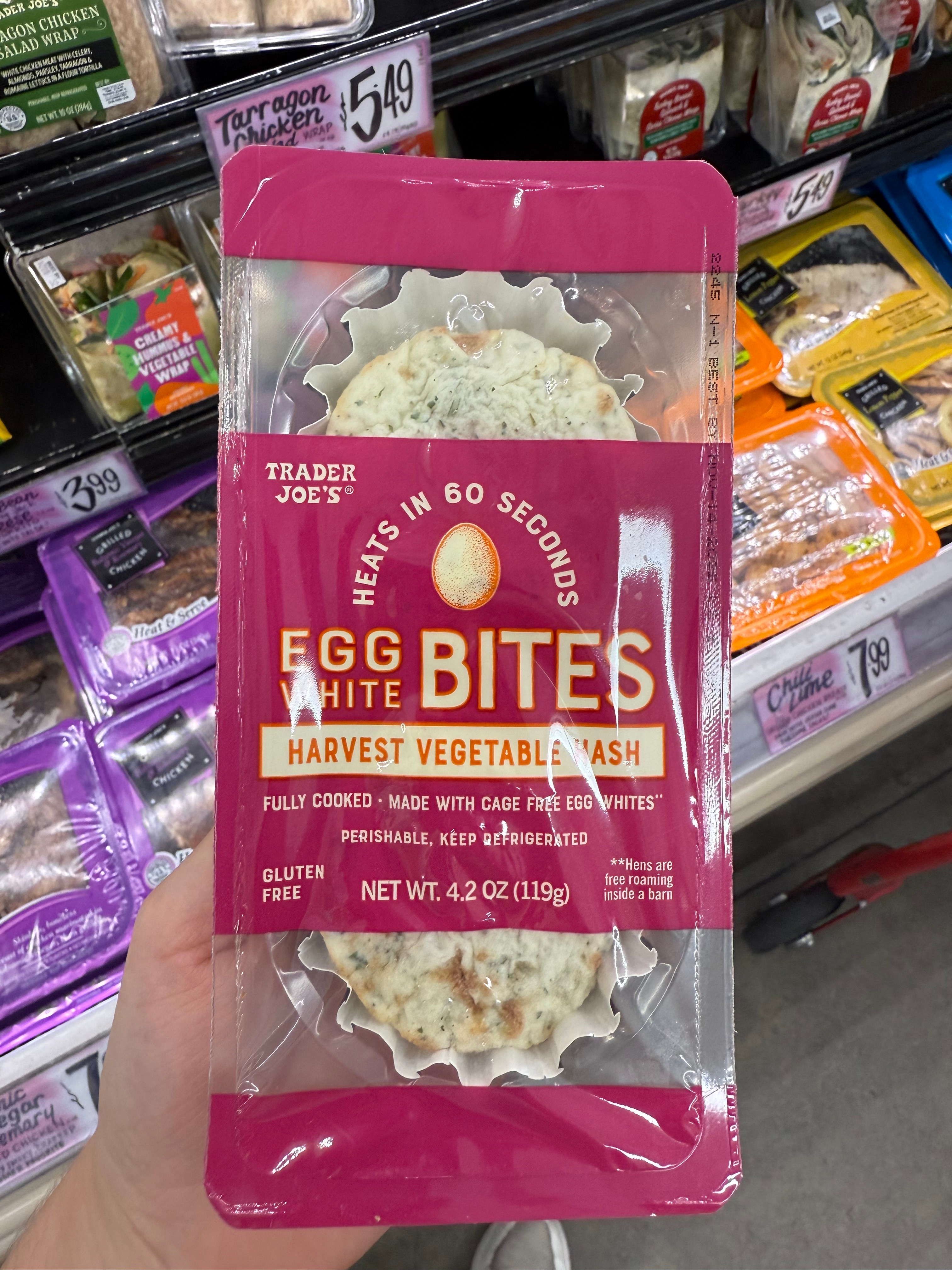 Hand holds Trader Joe's Egg White Bites package labeled "Harvest Vegetable," gluten-free, in a store display with other packaged foods