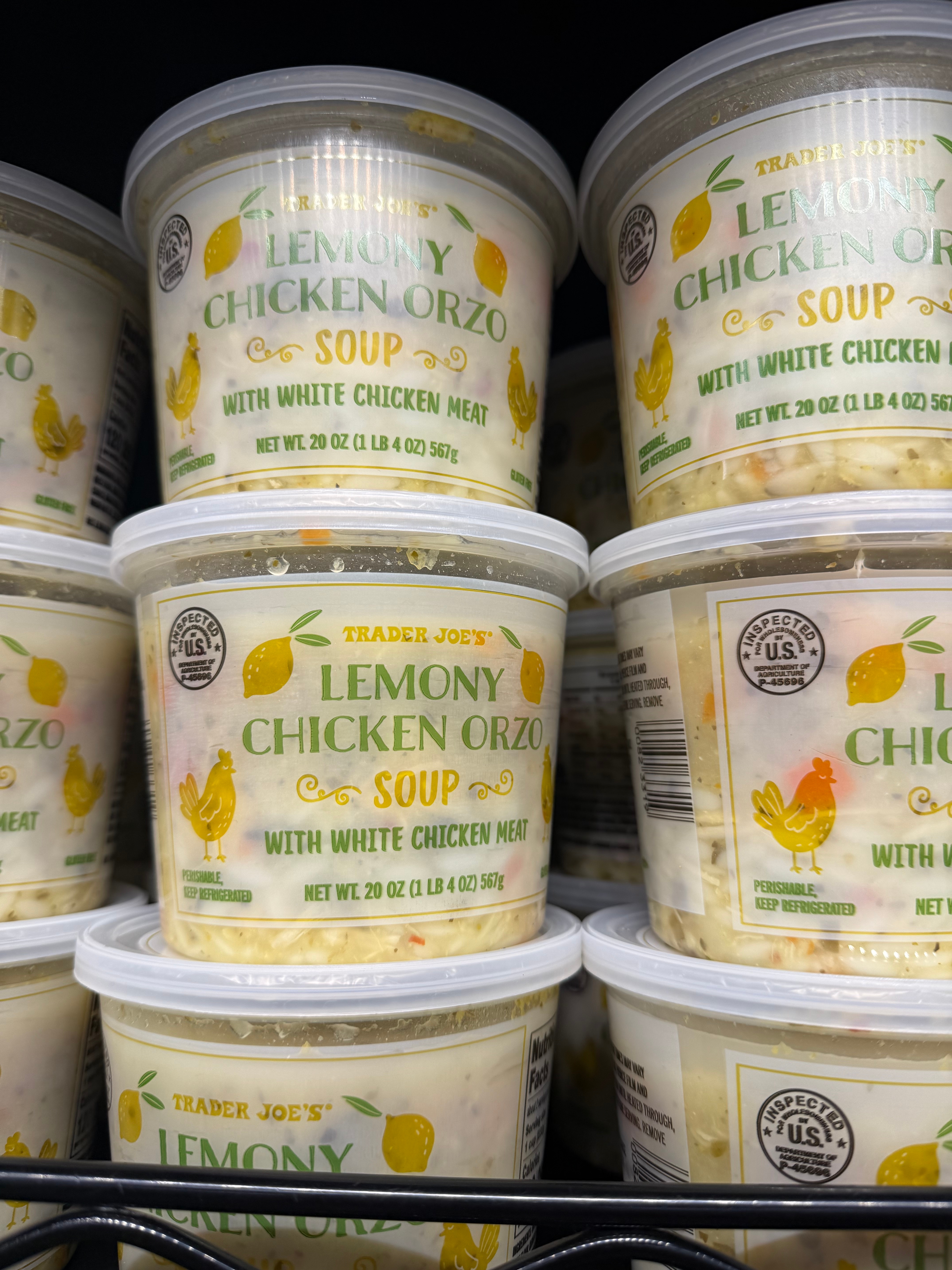 Trader Joe's Lemony Chicken Orzo Soup in 20 oz containers stacked on a shelf, labeled as containing white chicken meat