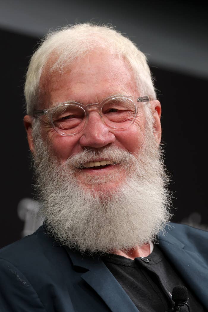 Smiling man with glasses and a full white beard, wearing a dark blazer over a black shirt at a formal event