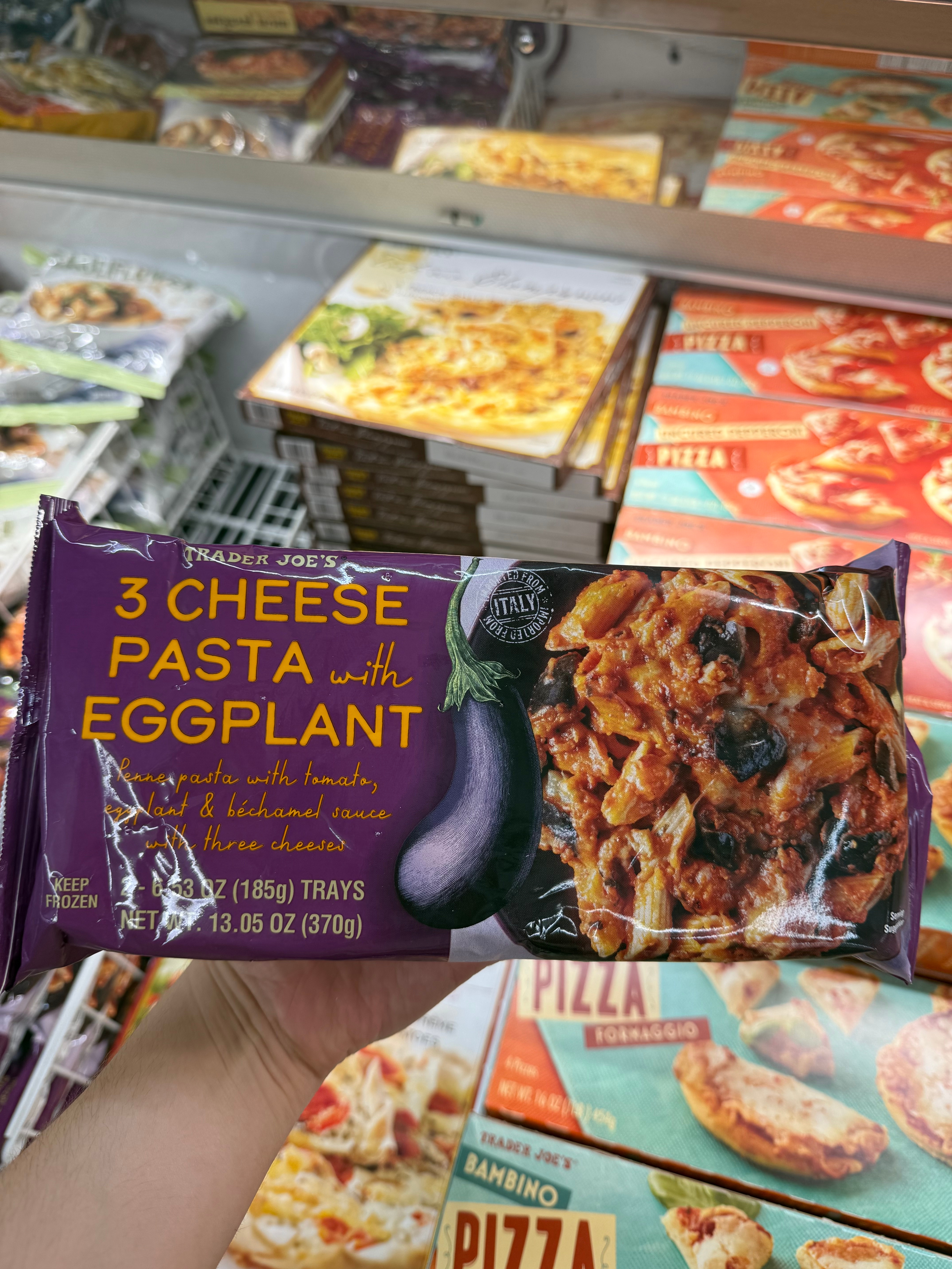 Package of 3 Cheese Pasta with Eggplant being held at a store. Text highlights tomatoes, eggplant, and Bechamel sauce ingredients. Pizza boxes visible behind