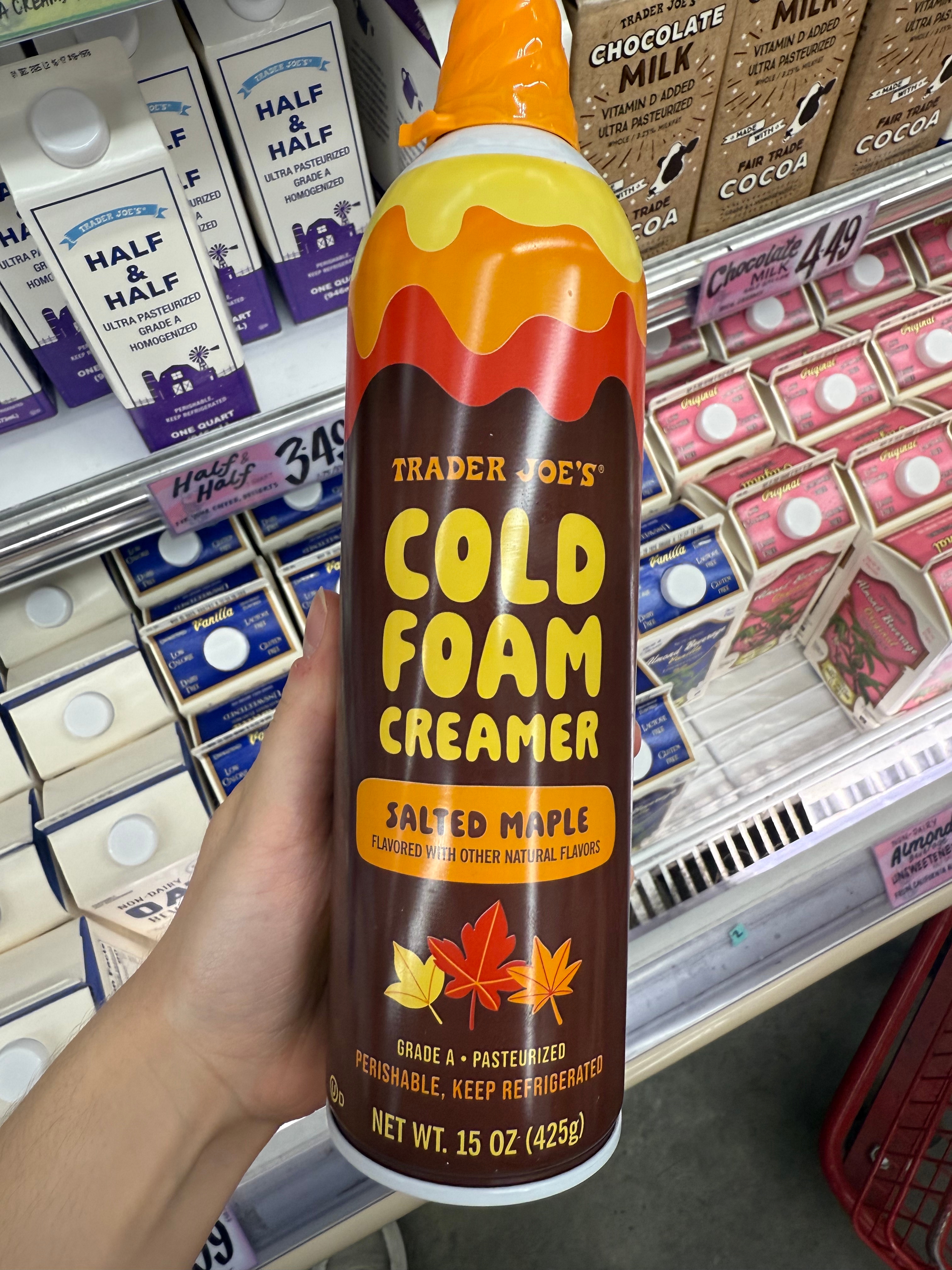 A hand holds a can of Trader Joe's Cold Foam Creamer, Salted Maple flavor, in a grocery store dairy aisle