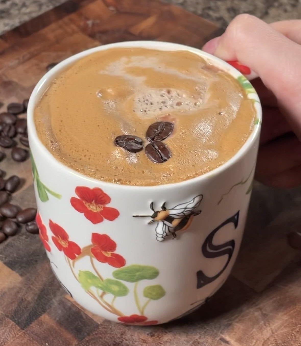 Coffee in a floral mug with foam topped with coffee beans, held by a hand on a wooden surface with scattered coffee beans